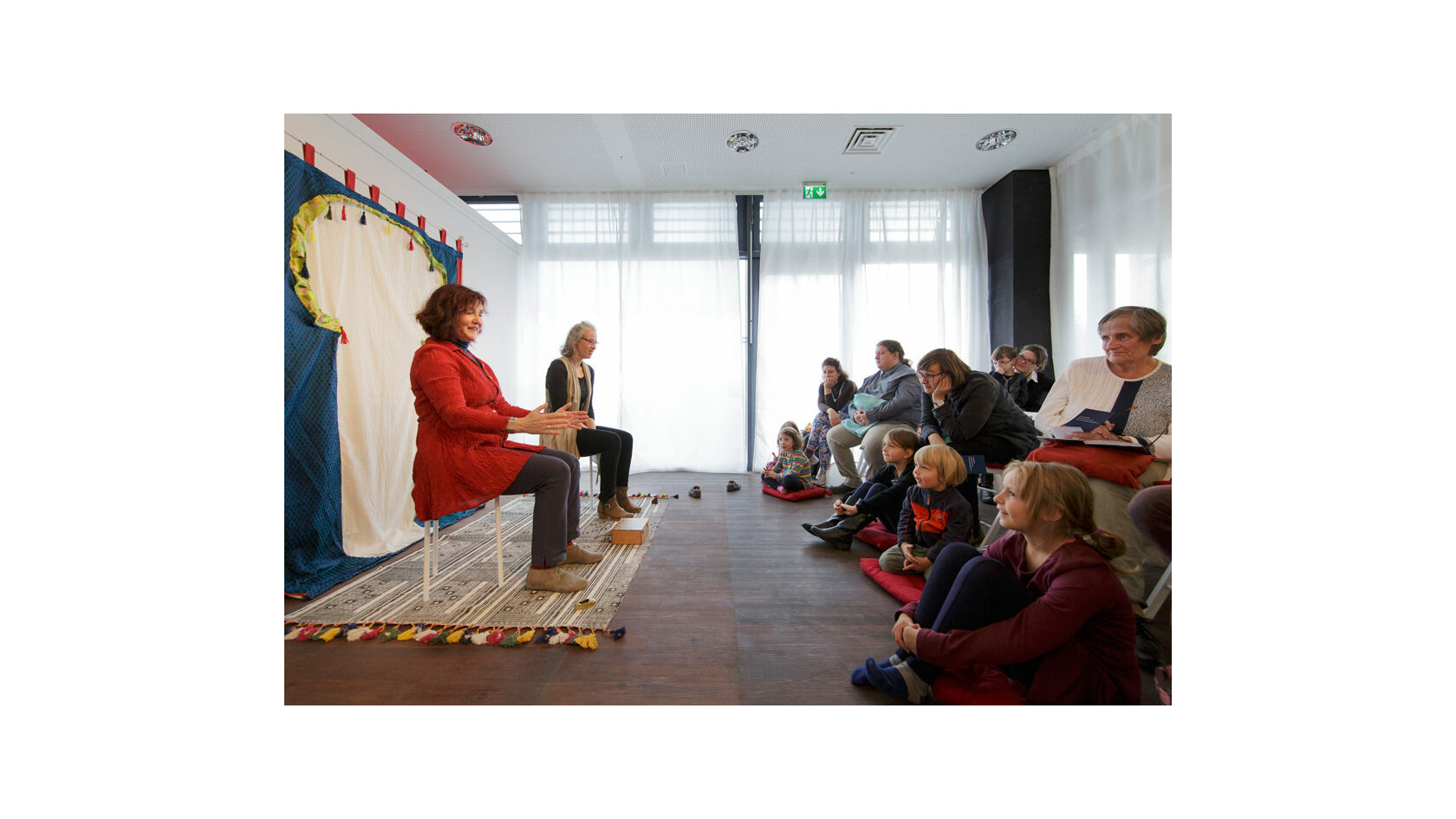 Brit Magdon vom Verein Erzählraum e. V. sitzt auf einem ausgerollten Teppich in einem Raum und erzählt vor Kindern und Erwachsenen, die ihr zuhören.